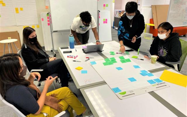 3 students and 2 instructors are discussing and placing colorful sticky notes on a large sticky note pad