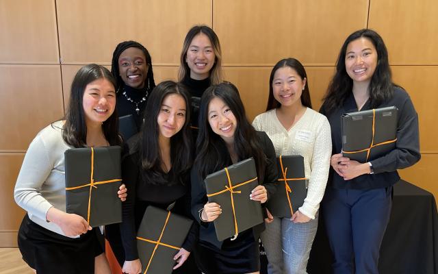 A group of smiling young women holding gifts wrapped in black and orange.