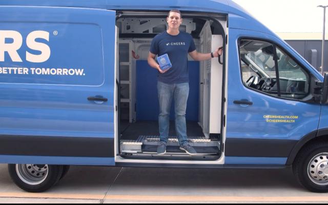 Young man in Blue Cheers t-shirt standing in the door of a blue delivery van with the Cheers logo on the side.