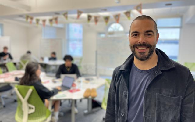 A smiling Ben Lehnert standing in the co-working space the the Princeton Entrepreneurial Hub - students are working in the background.